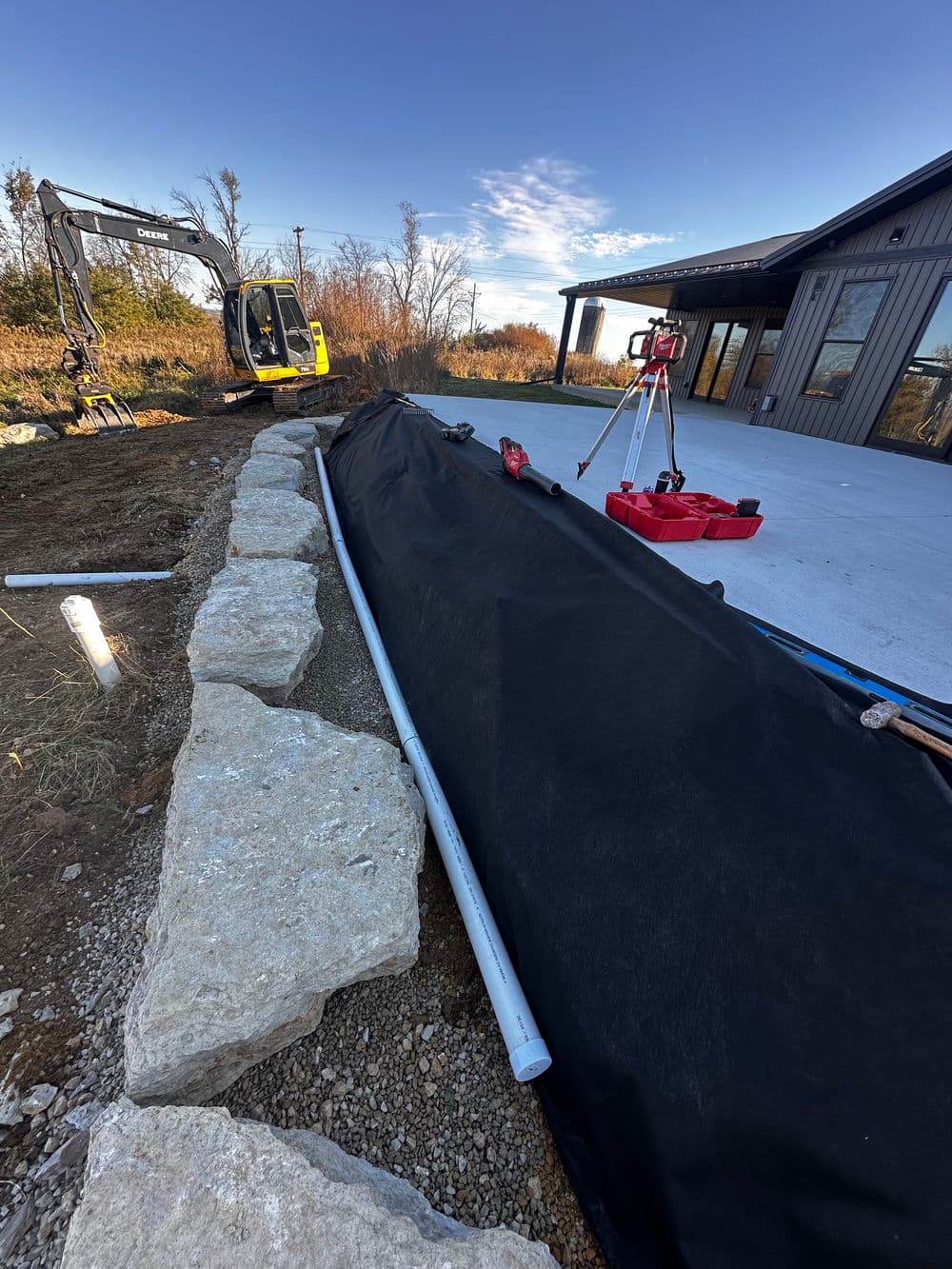 Excavation site with drainage pipe, landscaping fabric, and stone border near a newly constructed home.