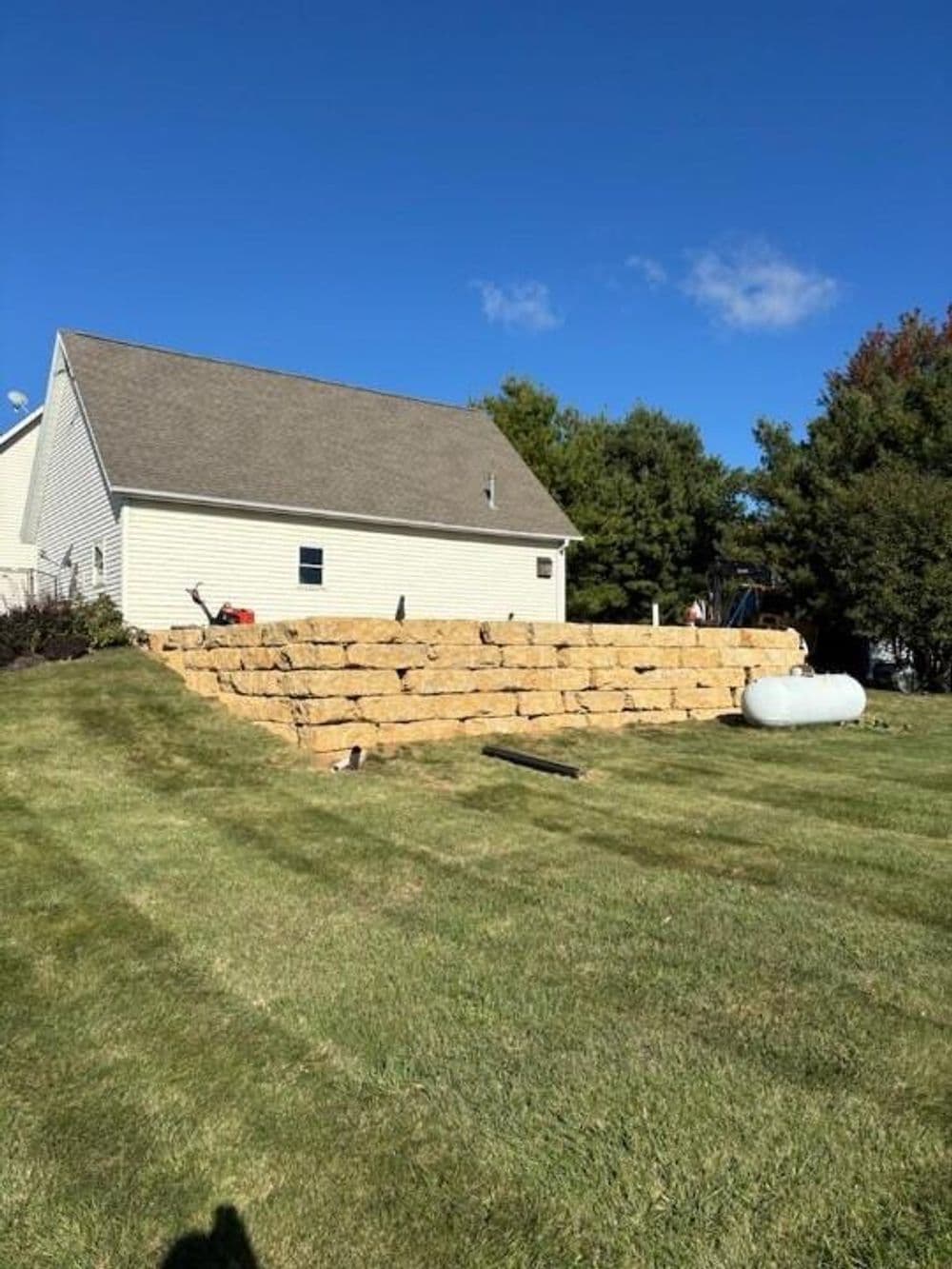 Home garden with large stacked stone retaining wall and lawn under blue sky.