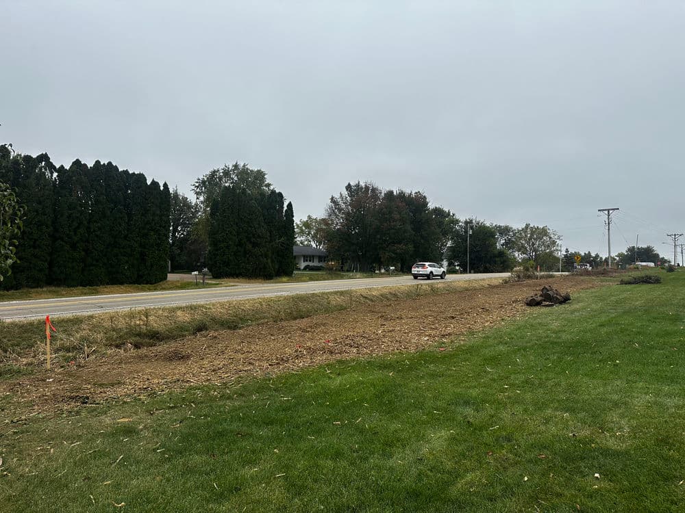 Car driving on a road beside a cleared field and trees on a cloudy day.