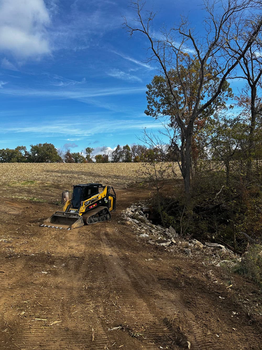 Skid steer loader clearing land near a rocky stream under a blue sky with scattered clouds.