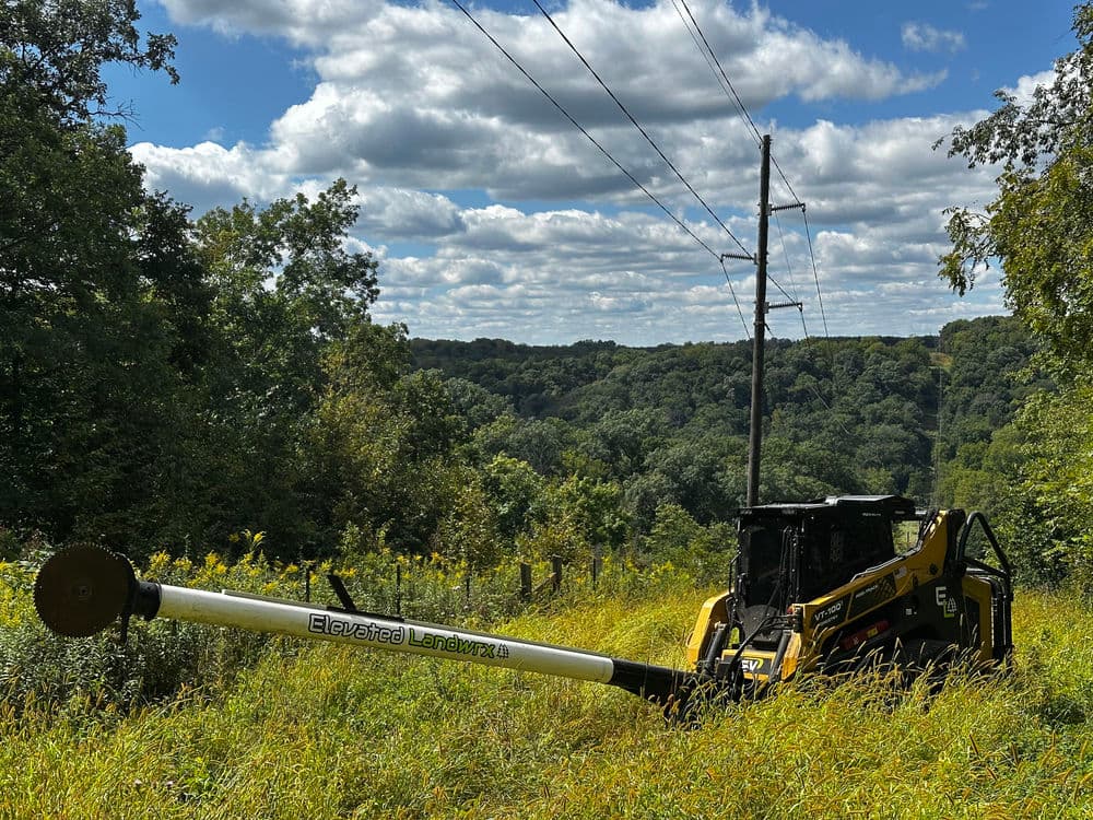 Heavy machinery working on a hillside, with power lines and a lush landscape in the background.