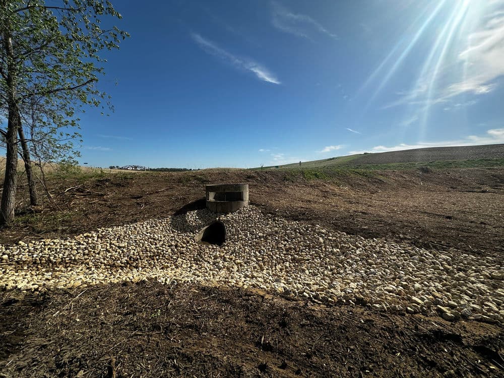 Stormwater drainage system with rocks on a clear sunny day, surrounded by green fields.