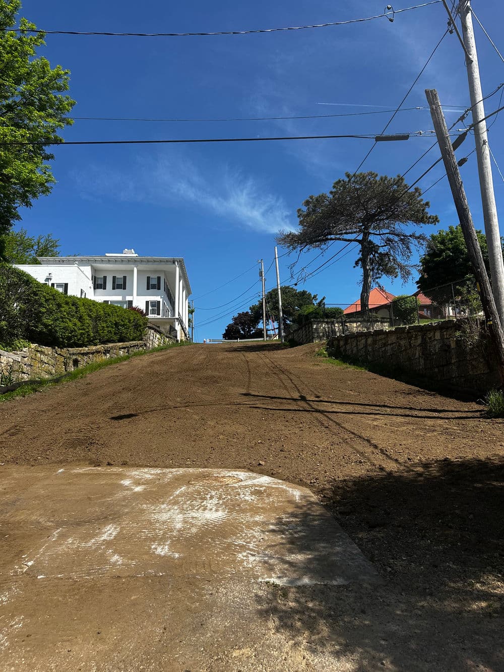 Dirt road leading uphill with utility poles, blue sky, and buildings in the background.