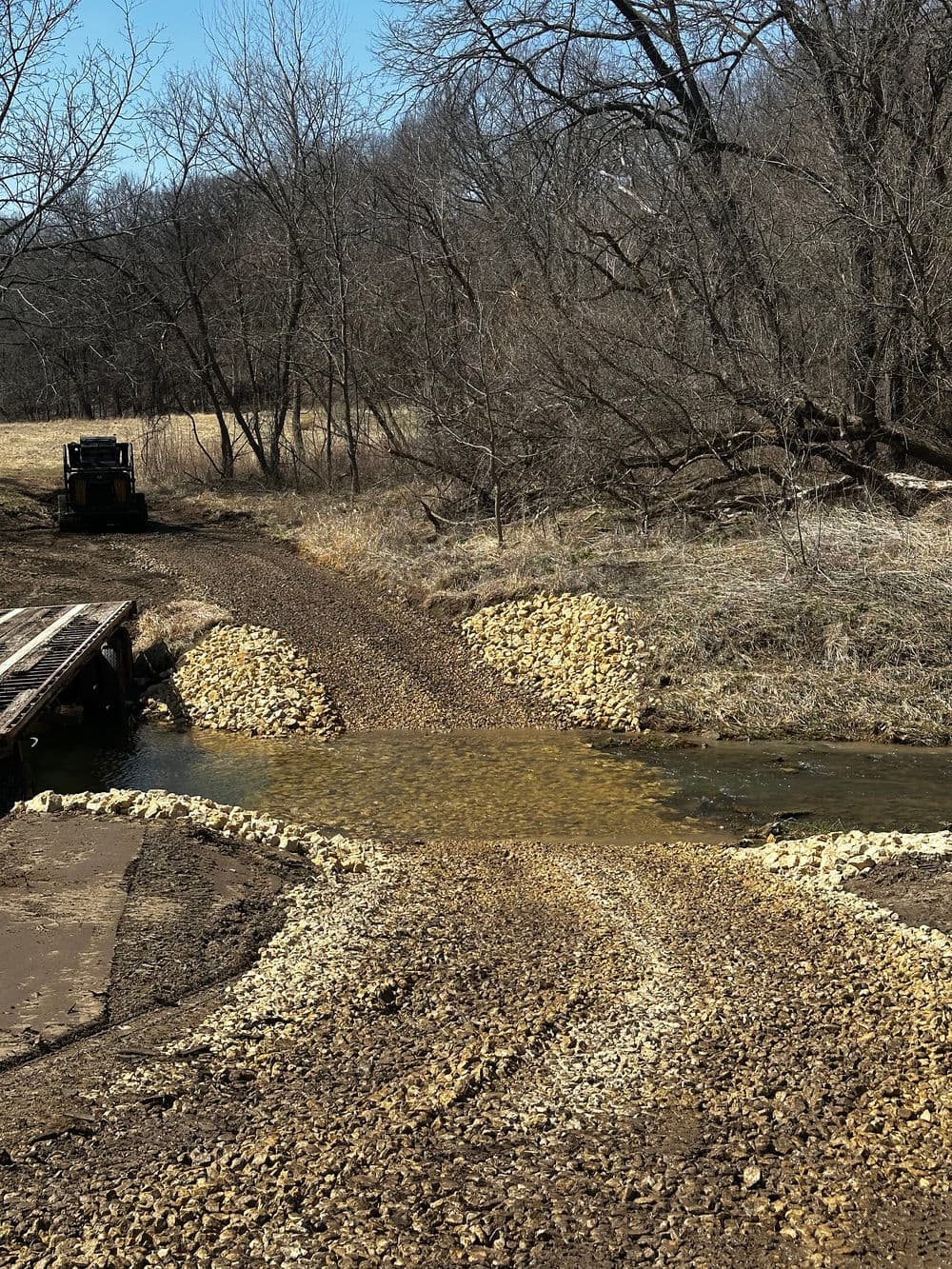 Gravel road crossing over a small stream with trees in the background, sunny day.