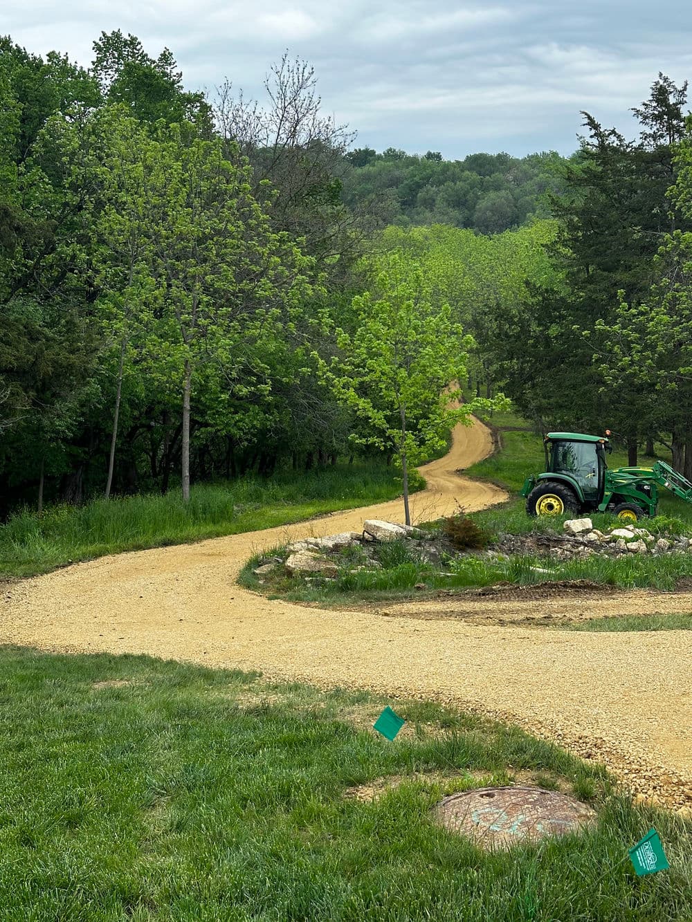 Tractor on winding gravel path through green landscape with trees and hills in background.