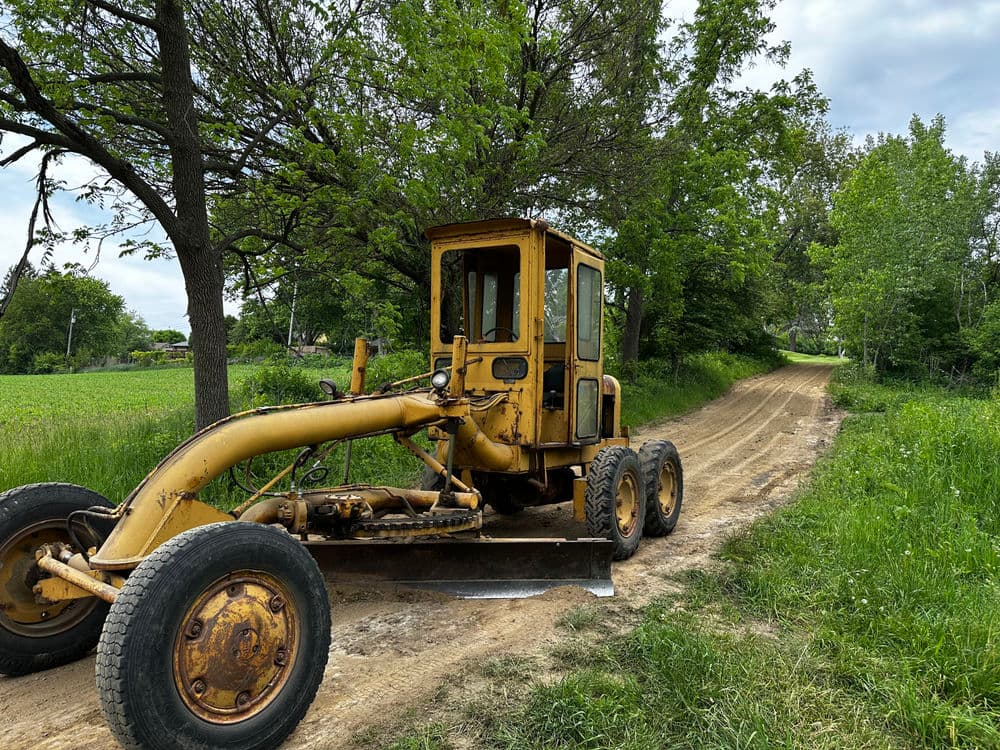Yellow grader on a dirt road surrounded by green trees and fields.
