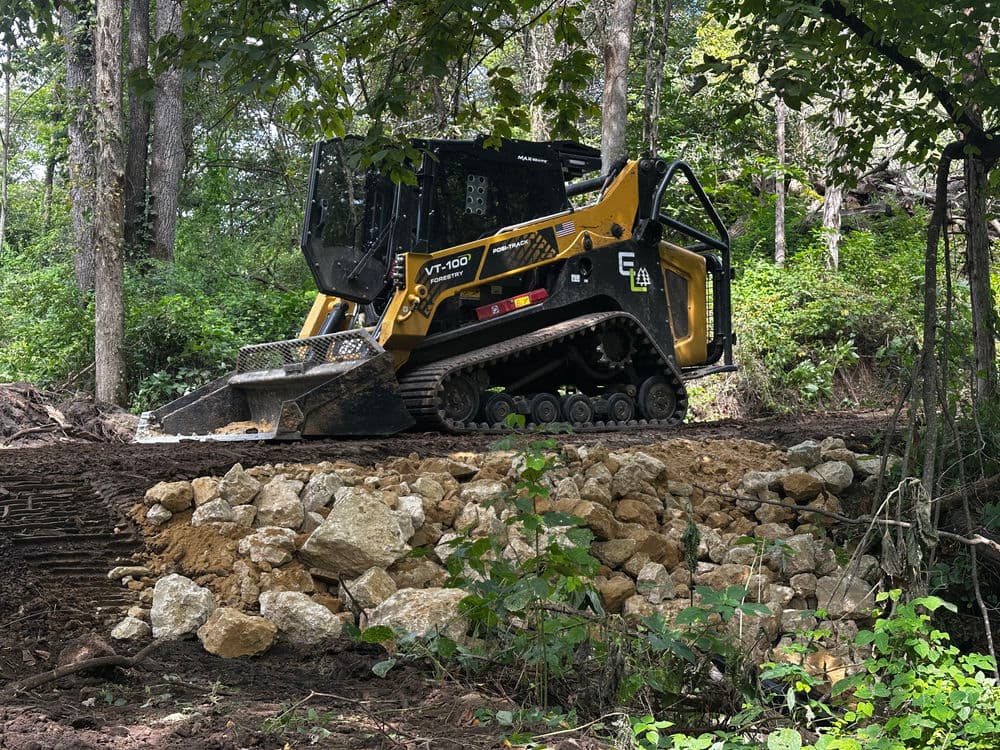 Yellow compact track loader working on a landscape project with rocks and trees in the background.